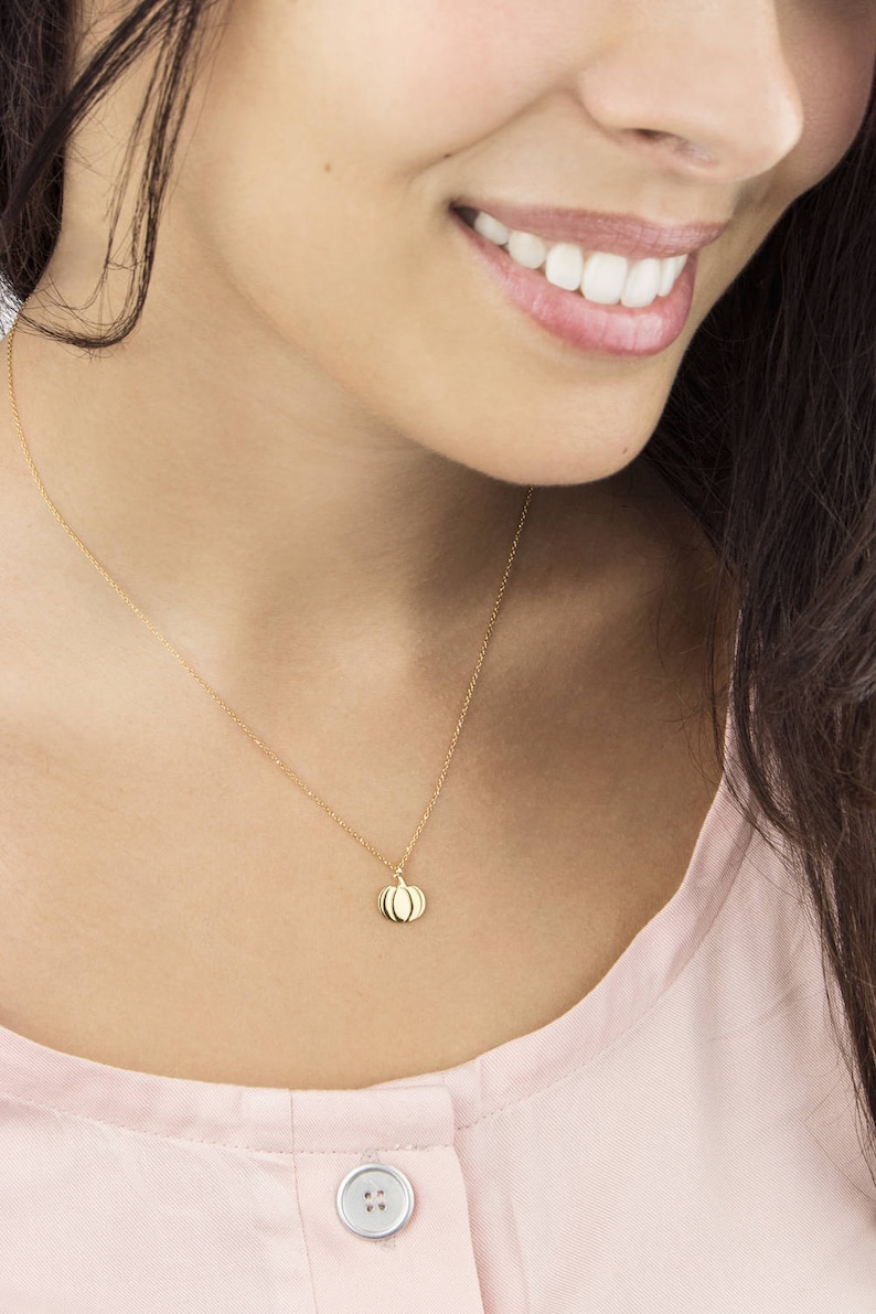 A yellow gold pendant necklace with a pumpkin charm with engraved details is shown worn by a model.