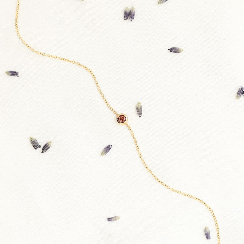 A special bracelet with a deep red garnet gemstone and an adjustable chain in yellow gold was laid down on a white background, surrounded by leaves.