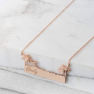 A rose gold pendant necklace with a bar that resembles a beach with an engravable message, a palm tree on each side an umbrella on the left, and the sun on the right is shown on a marble surface.