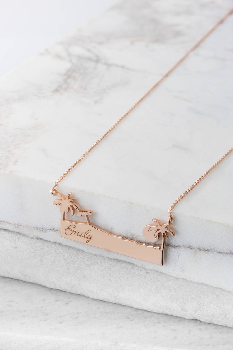 A rose gold pendant necklace with a bar that resembles a beach with an engravable message, a palm tree on each side an umbrella on the left, and the sun on the right is shown on a marble surface.