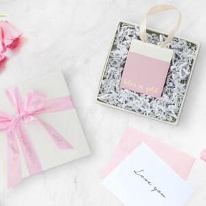 A beautiful picture displaying the packaging that the bracelet comes in with flowers on each side, all laid down on a marble floor. A white box with a pink ribbon and a little bag in pink and white that contains the item.