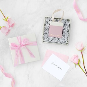 A beautiful picture displaying the packaging that the bracelet comes in with flowers on each side, all laid down on a marble floor. A white box with a pink ribbon and a little bag in pink and white that contains the item.