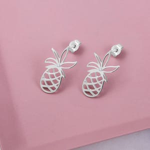 A delicate white gold pineapple stud earring with a white natural diamond in the center of the leaves is shown on a pink surface.