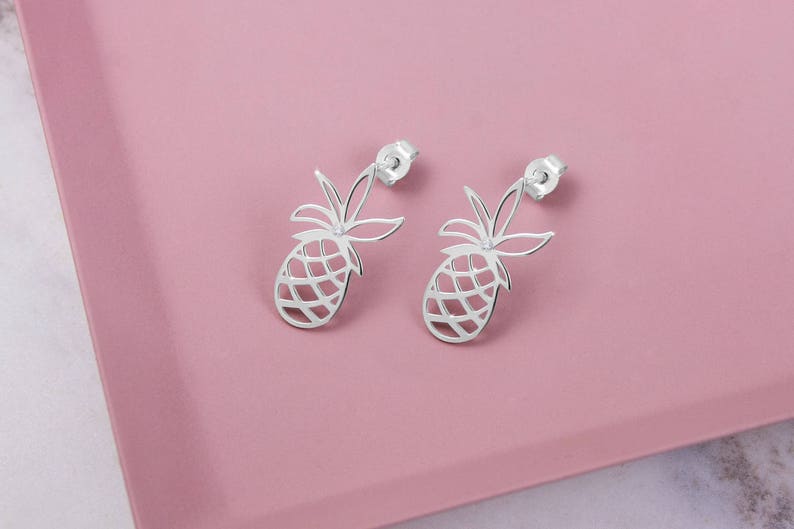 A delicate white gold pineapple stud earring with a white natural diamond in the center of the leaves is shown on a pink surface.