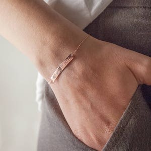 A rose gold bracelet with an engravable horizontal curved bar with coordinates attached to a chain that has its jump rings soldered and a spring-ring clasp is shown worn by a model.