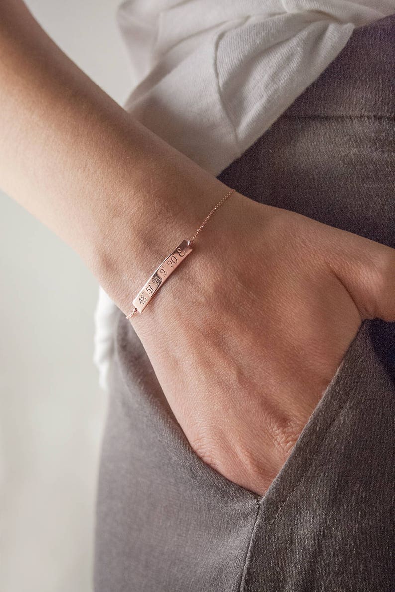 A rose gold bracelet with an engravable horizontal curved bar with coordinates attached to a chain that has its jump rings soldered and a spring-ring clasp is shown worn by a model.