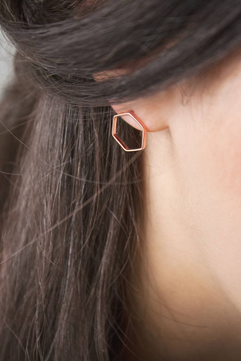 A thin hexagon-shaped stud earring in rose gold and with a push-back closure is shown worn by a model.