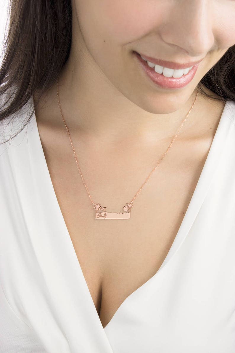 A rose gold pendant necklace with a bar that resembles a beach with an engravable message, a palm tree on each side an umbrella on the left, and the sun on the right is shown worn by a model.