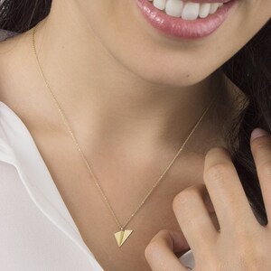 Op de afbeelding: Een gouden ketting met een klein papieren vliegtuigje als hanger. De ketting wordt gedragen door iemand met een wit shirt.
