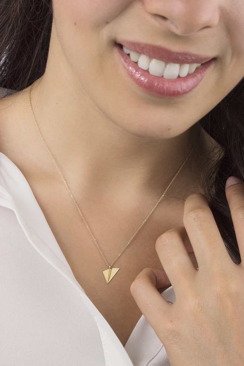 Op de afbeelding: Een gouden ketting met een klein papieren vliegtuigje als hanger. De ketting wordt gedragen door iemand met een wit shirt.