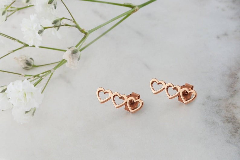 A pair of three-heart outline stud earrings in rose gold is shown on a marble surface surrounded by flowers.