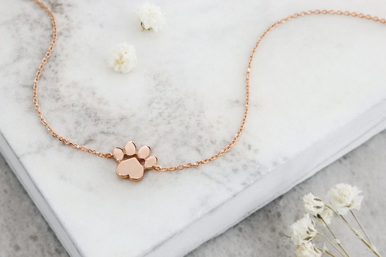 A paw charm bracelet with a distinct separation between the pads and an adjustable chain in rose gold laid down a piece of marble with pedals spread across the picture