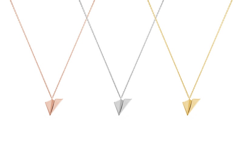 All three color options of the solid gold necklace with a tiny flat paper plane charm attached to it. Rose, white and yellow gold, every option displayed right next to each other on a white background.