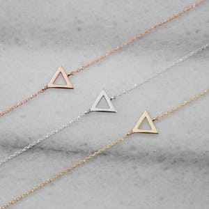 All three color options of hand-crafted bracelet featuring a triangle frame charm in yellow, white and rose gold. A minimalist style piece with an adjustable chain laid down on a marble piece.