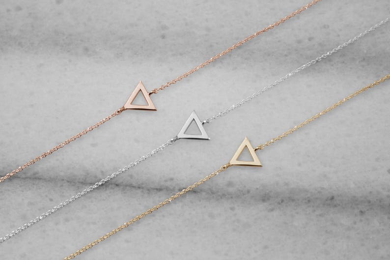 All three color options of hand-crafted bracelet featuring a triangle frame charm in yellow, white and rose gold. A minimalist style piece with an adjustable chain laid down on a marble piece.