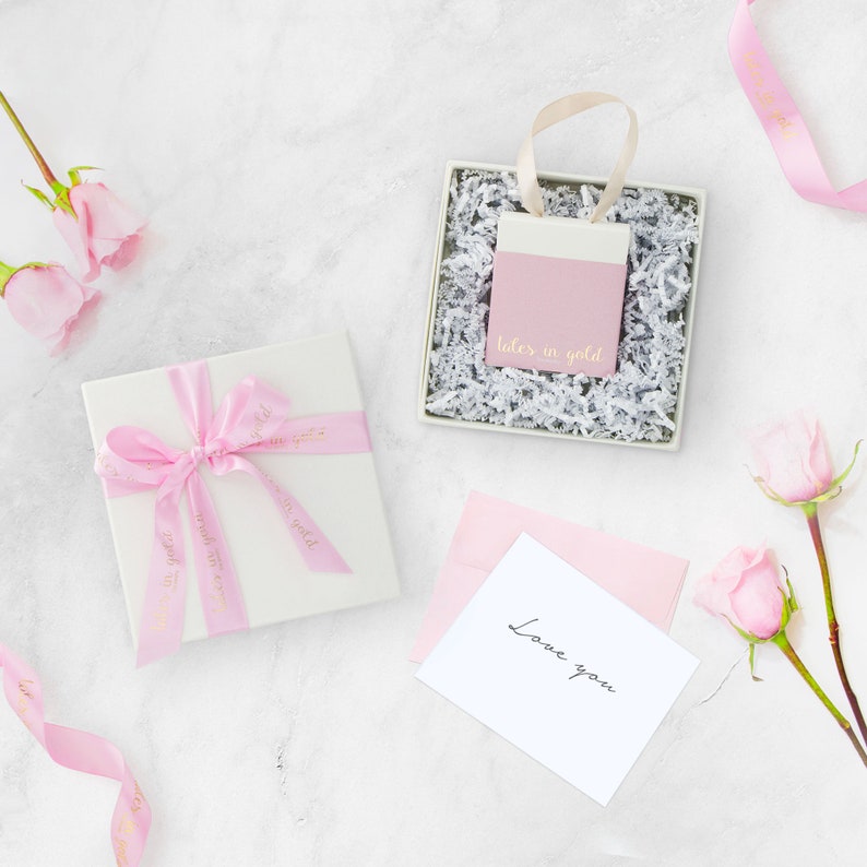 A beautiful picture displaying the packaging that the bracelet comes in with flowers on each side, all laid down on a marble floor. A white box with a pink ribbon and a little bag in pink and white that contains the item.