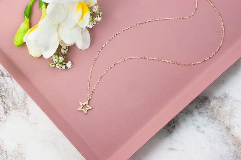 Laid down on a pink display piece and with flowers next to it, a yellow gold necklace with a special star outline charm and natural white diamonds embedded in it.