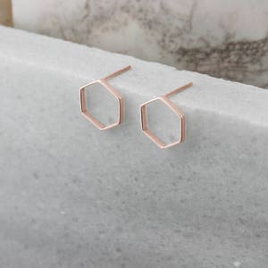 A pair of thin hexagon-shaped stud earrings in rose gold and with a push-back closure is shown on a marble surface with flowers surrounding it.