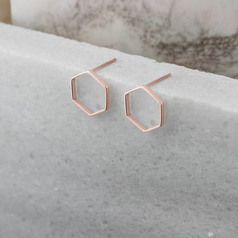 A pair of thin hexagon-shaped stud earrings in rose gold and with a push-back closure is shown on a marble surface with flowers surrounding it.