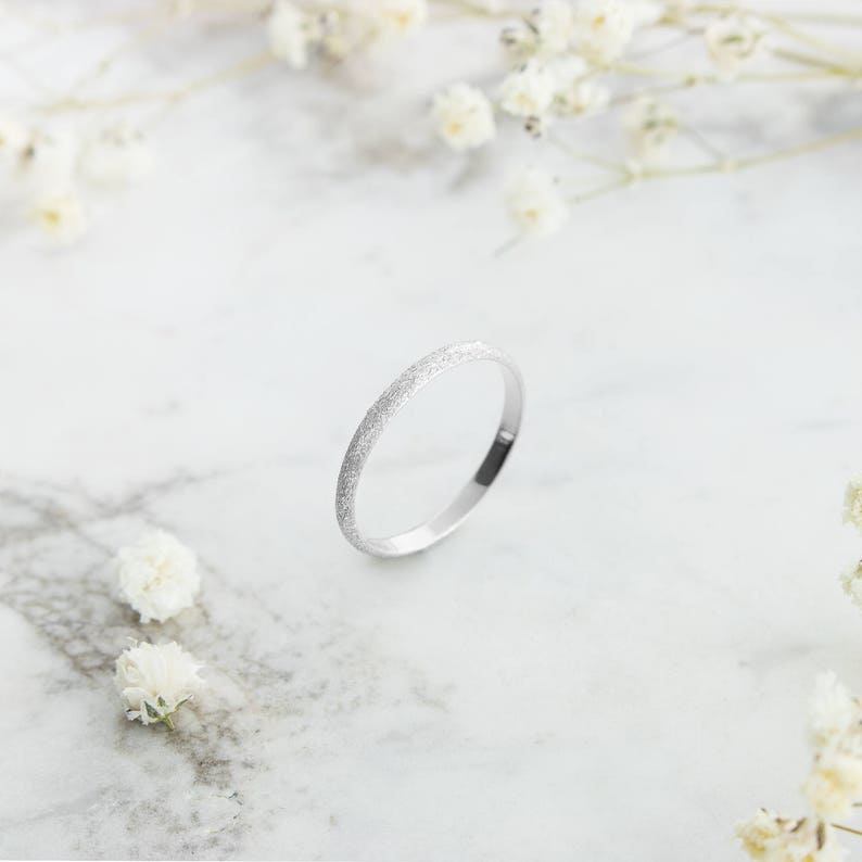 A classic and unique white gold band ring with a sandstone finish laid down on a marble display surface, surrounded by flowers.