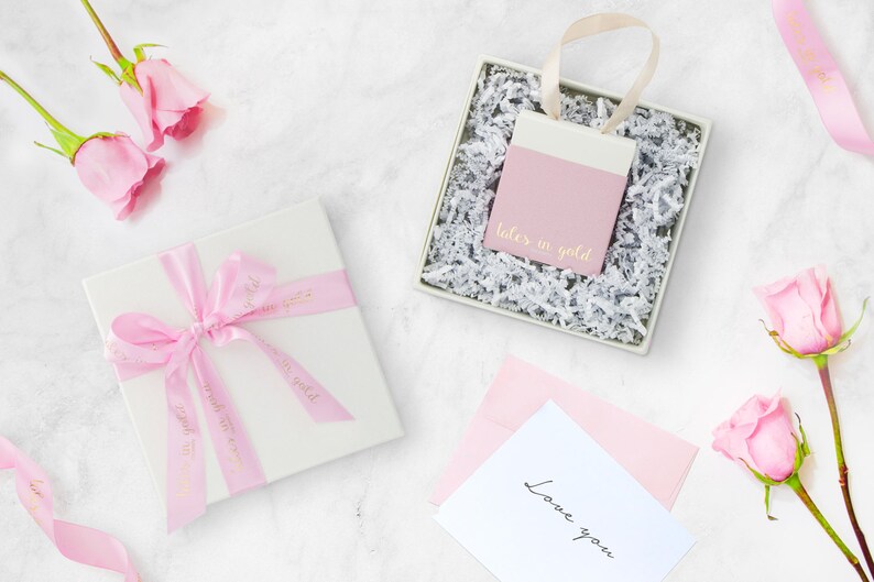 A beautiful picture displaying the packaging that the bracelet comes in with flowers on each side, all laid down on a marble floor. A white box with a pink ribbon and a little bag in pink and white that contains the item.