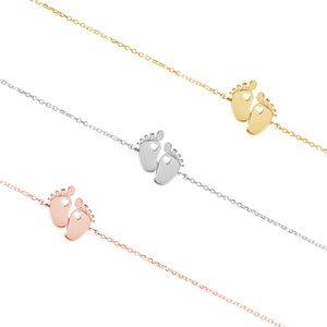 All three color options of the tiny gold baby feet charm with a heart in each one of the feet and an adjustable chain surrounding it. Yellow, white and rose gold, all displayed on a white background right next to each other.