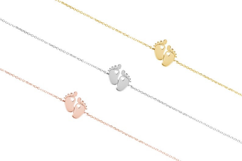 All three color options of the tiny gold baby feet charm with a heart in each one of the feet and an adjustable chain surrounding it. Yellow, white and rose gold, all displayed on a white background right next to each other.