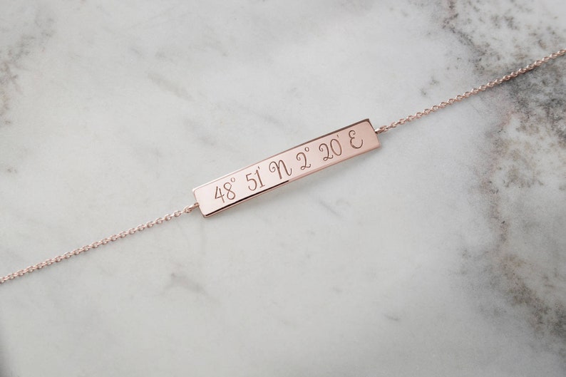 A rose gold bracelet with an engravable horizontal curved bar with coordinates attached to a chain that has its jump rings soldered and a spring-ring clasp is shown on a marble surface.