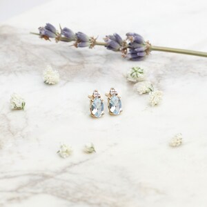 A unique pair of stud earrings in yellow gold with a light sky blue topaz gemstone and a round white natural diamond worn by a model.