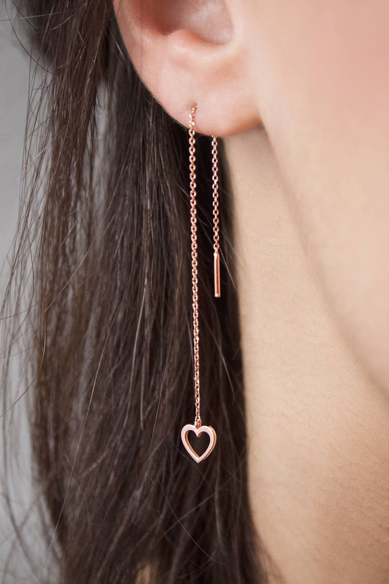 Worn by a model, A threader earring with a dangling heart outline on one end in rose gold.