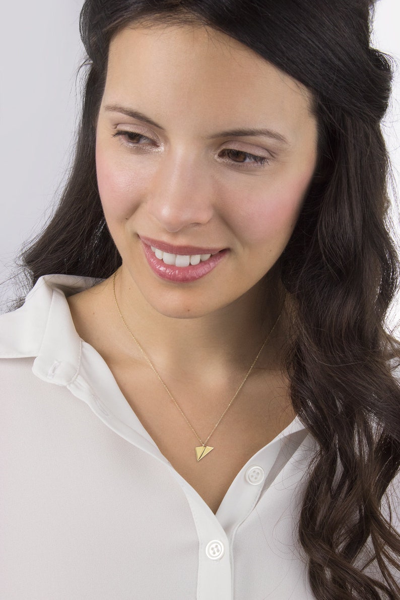 A unique yellow gold necklace with a tiny flat paper plane charm attached to it is shown worn by a model.