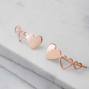 A pair of stunning rose gold and different sized three heart climber earrings with the biggest of the hearts being solid and the other two have just the outline is shown worn by a woman.