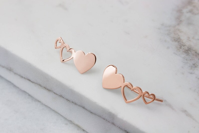 A pair of stunning rose gold and different sized three heart climber earrings with the biggest of the hearts being solid and the other two have just the outline is shown worn by a woman.