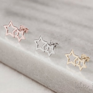 May include: Three pairs of star-shaped earrings in gold, silver, and rose gold. The earrings are made of two stars, one slightly larger than the other, with the smaller star overlapping the larger star. The earrings are on a white marble surface.