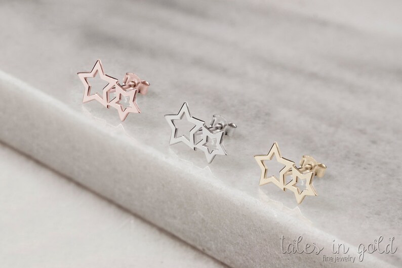 May include: Three pairs of star-shaped earrings in gold, silver, and rose gold. The earrings are made of two stars, one slightly larger than the other, with the smaller star overlapping the larger star. The earrings are on a white marble surface.