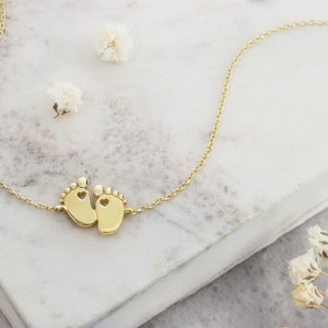 A tiny yellow gold baby feet charm with a heart in each one of the feet and an adjustable chain surrounding it, laid down on a marble piece with feathers surrounding it.