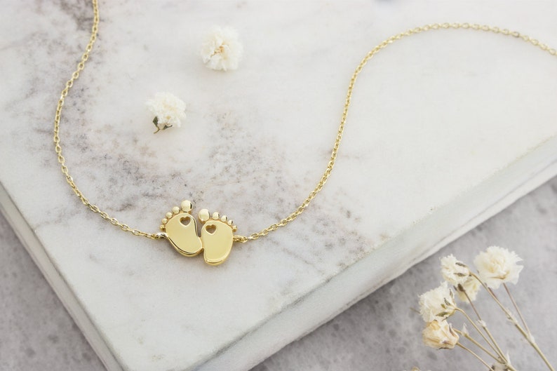 A tiny yellow gold baby feet charm with a heart in each one of the feet and an adjustable chain surrounding it, laid down on a marble piece with feathers surrounding it.