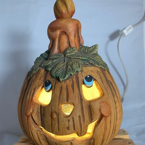 May include: A ceramic pumpkin shaped light with a lit candle on top. The pumpkin has a carved face with blue eyes and a wide smile. The pumpkin is brown with a textured surface.