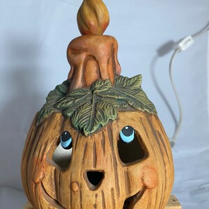 May include: A ceramic pumpkin candle holder with a smiling face, painted blue eyes, and a candle on top. The pumpkin is brown with green leaves around the candle.