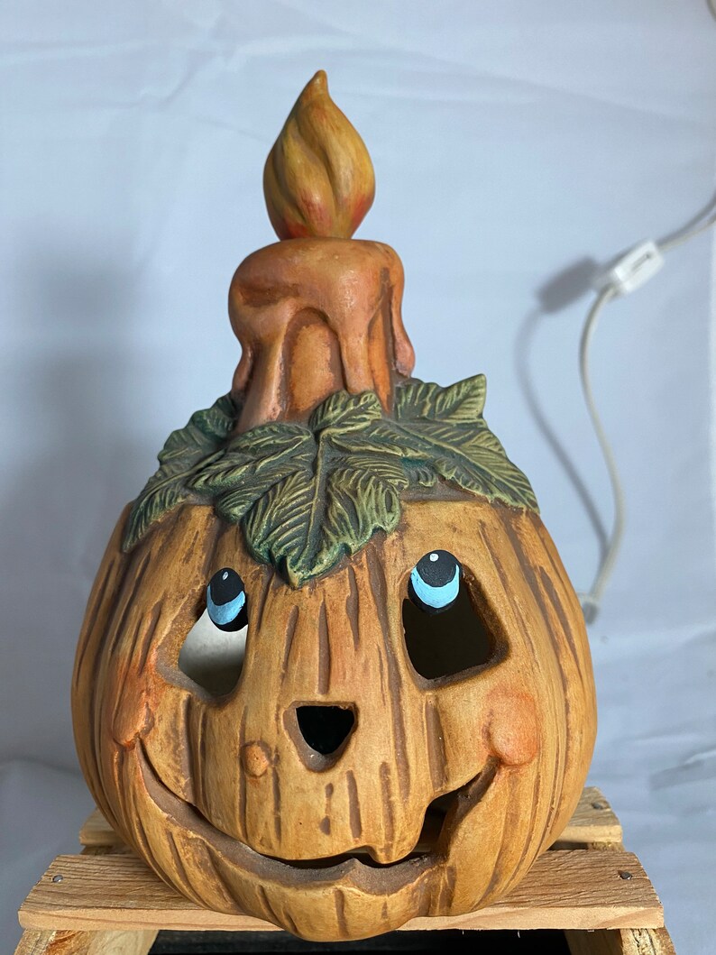 May include: A ceramic pumpkin candle holder with a smiling face, painted blue eyes, and a candle on top. The pumpkin is brown with green leaves around the candle.