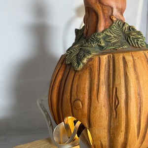 May include: A ceramic pumpkin lamp with a brown and green finish. The lamp has a light bulb inside and a cord coming out of the bottom. The pumpkin is sitting on a wooden base.