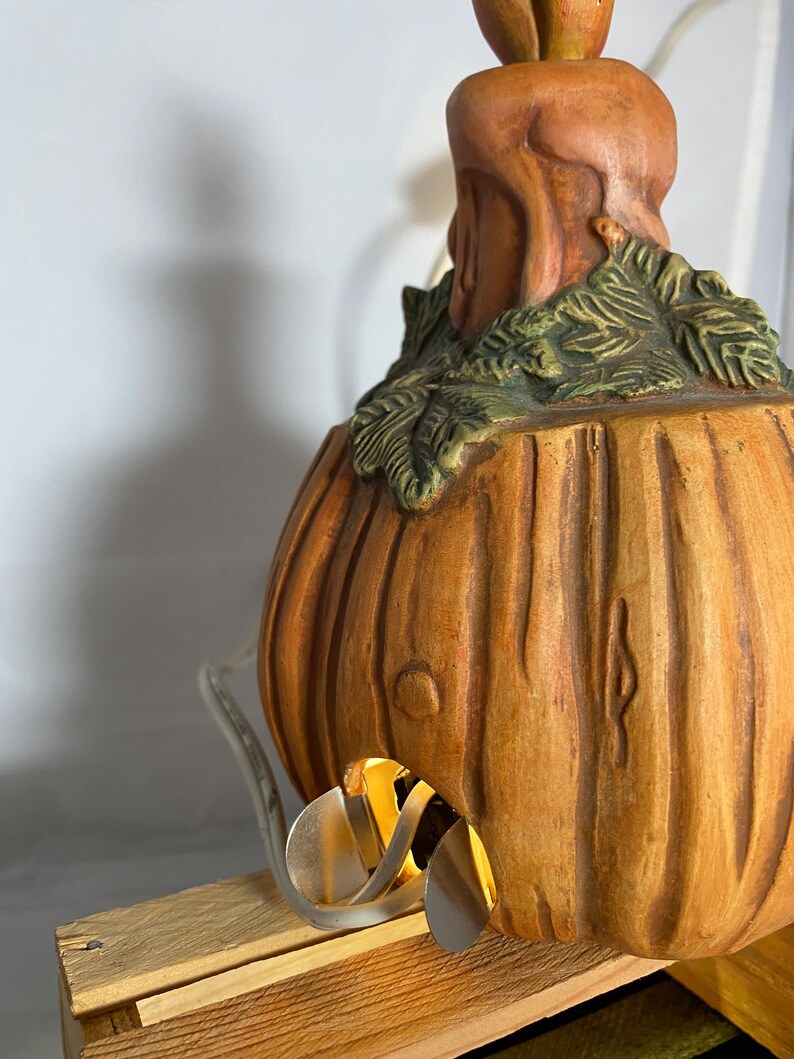 May include: A ceramic pumpkin lamp with a brown and green finish. The lamp has a light bulb inside and a cord coming out of the bottom. The pumpkin is sitting on a wooden base.
