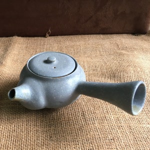 Puede incluir: Una tetera de cerámica gris con tapa y un pico largo, y un tarro con tapa de cerámica gris a juego.