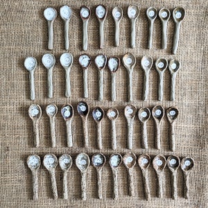 May include: A collection of 80 small ceramic spoons with white numbers on them. The spoons are arranged in rows on a hessian background.