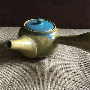 Puede incluir: Una tetera de cerámica amarilla y azul con un pico largo y una tapa. La tetera está sobre una superficie de arpillera.