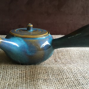 May include: A teal blue ceramic teapot with a brown lid and a long, thin handle. The teapot has a glossy finish and is sitting on a hessian surface.