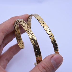 May include: Two gold-coloured metal bracelets. One bracelet has a braided design, and the other has a twisted design.