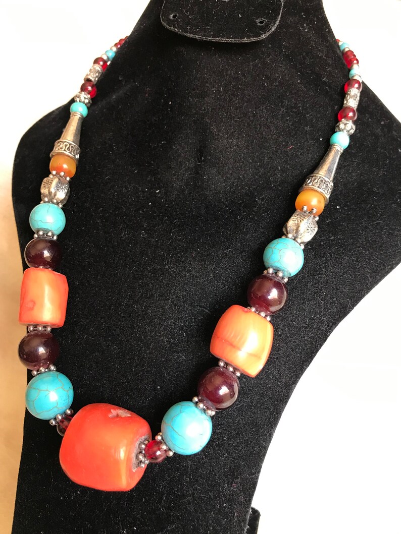K&ouml;nnte beinhalten: Eine mehrfarbige Perlenkette mit orangefarbenen, t&uuml;rkisfarbenen und roten Perlen. Die Kette hat silberne Akzente und wird auf einem schwarzen Samtst&auml;nder pr&auml;sentiert.