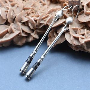 May include: A pair of silver dangle earrings with black accents. The earrings are long and slender, with a small round bead at the top. The earrings are hanging from a piece of brown rock.