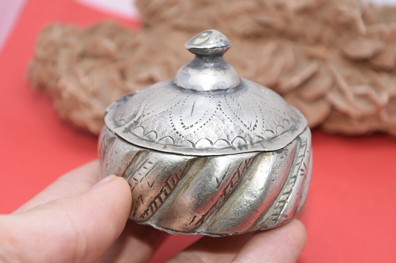 May include: A silver trinket box with an ornate, engraved lid and a twisted, textured base. The box is small and has a domed lid.
